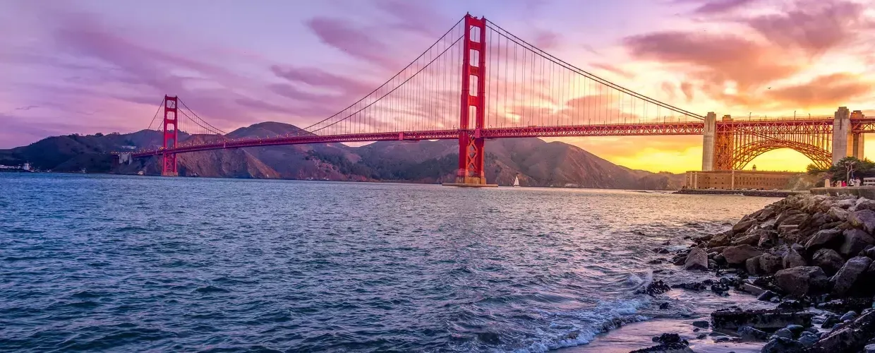 The Golden Gate Bridge at sunset with a multicolored sky and the San Francisco Bay in the foreground.